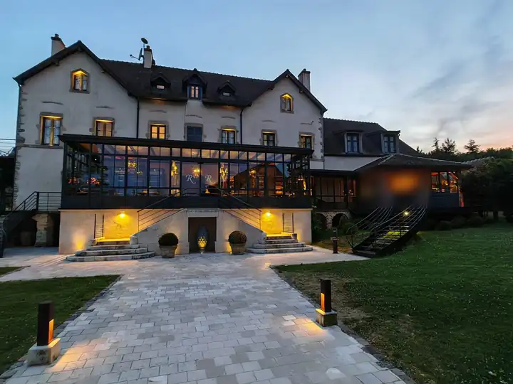 Herrenhaus des Domaine de Rymska am Abend mit beleuchtetem Wintergarten aus schwarzem Stahl und Glas, davor ein Weg aus hellem Pflaster