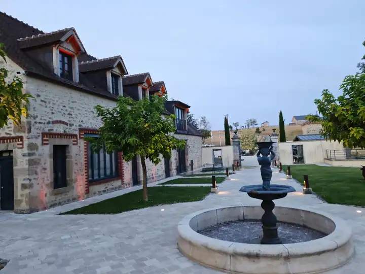 Hof mit rundem Brunnen und Gusseisenfigur vor Nebengebäuden aus Bruchstein am Abend