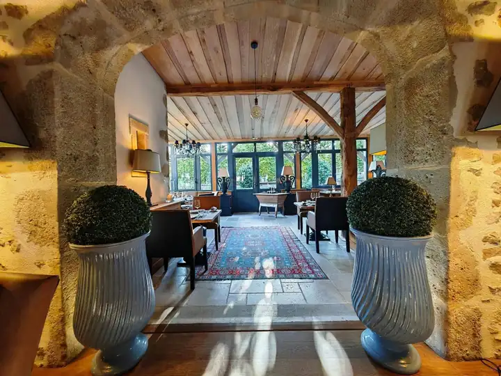 Blick durch einen Bogen aus Naturstein in den Speiseraum mit Holzdecke, Kronleuchtern und blauem Teppich, vorn zwei Buchsbaumkugeln in Vasen
