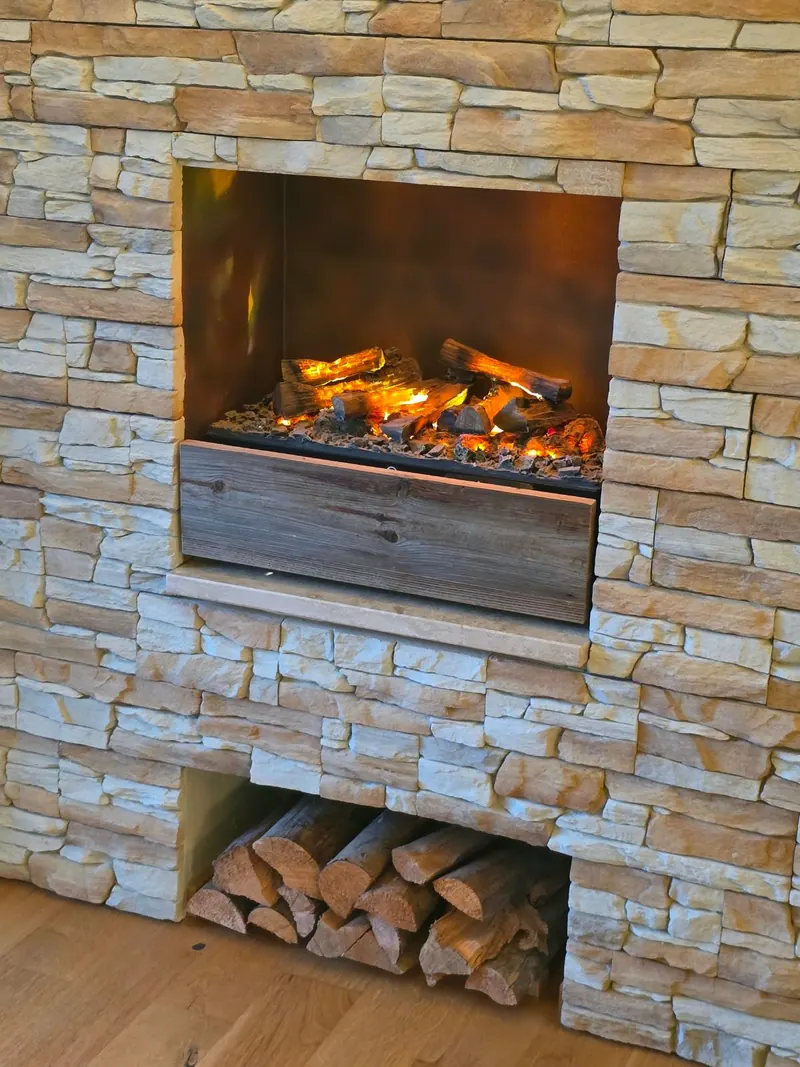 Kamin mit Flammen in einer Wand aus hellem Naturstein, darunter gestapeltes Brennholz