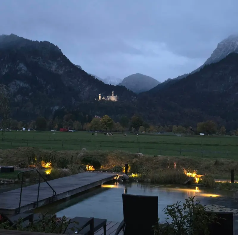 Abendblick aus dem Garten des Hotels Das Rübezahl: beleuchteter Naturschwimmteich mit Holzsteg, dahinter Wiesen und das angestrahlte Schloss Neuschwanstein vor den Bergen