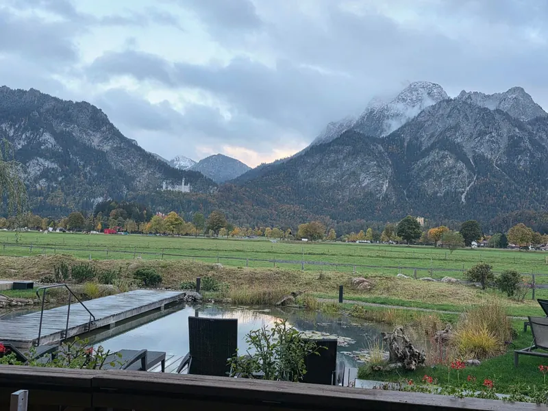 Blick über Garten, Teich und Wiesen auf Schloss Neuschwanstein und schneebedeckte Gipfel unter Wolken