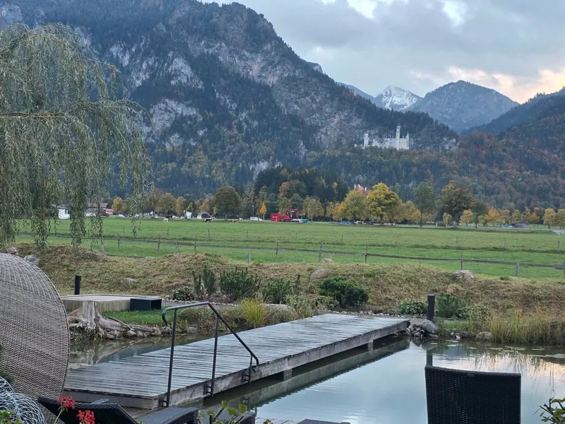 Holzsteg über einem Naturschwimmteich, dahinter eine weite Wiese, Wald und Schloss Neuschwanstein vor den Bergen