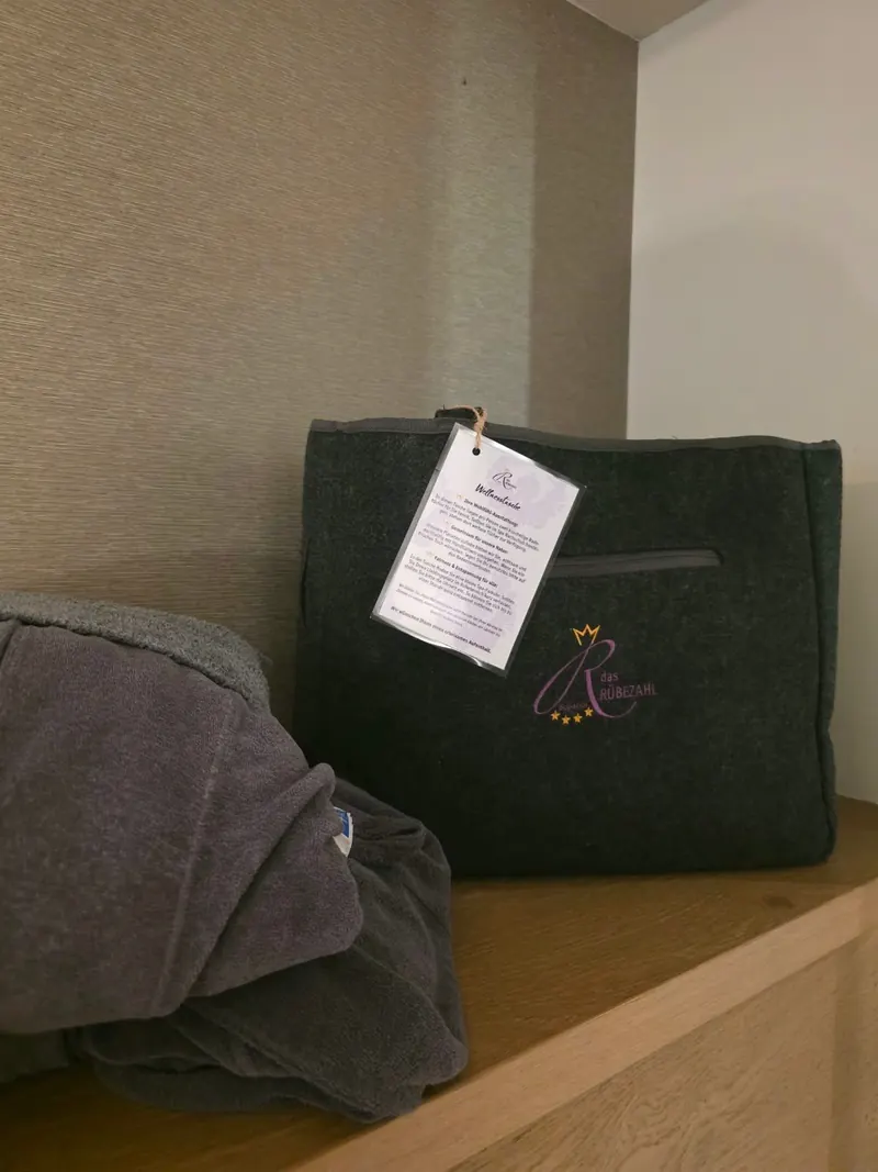 Graue Wellnesstasche aus Filz mit dem Logo des Rübezahl und einem Anhänger mit Hinweisen, daneben ein Bademantel