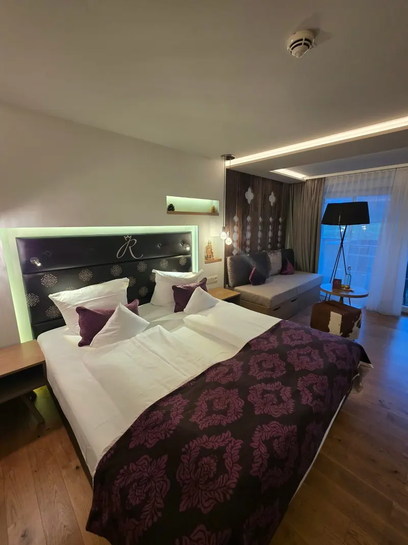 Hotelzimmer mit Doppelbett, weinrotem Damastüberwurf und lila Kissen, beleuchtetes Kopfteil, Sitzecke am Fenster
