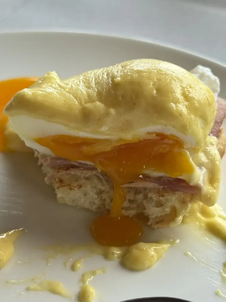 Angeschnittene Eggs Benedict mit Schinken auf Brot, darüber Sauce Hollandaise, das Eigelb läuft auf den weißen Teller