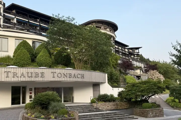 Eingang der Traube Tonbach mit dem Schriftzug über der Tür, Treppen und Beeten, darüber das Hotel mit dunklen Holzbalkonen am bewaldeten Hang