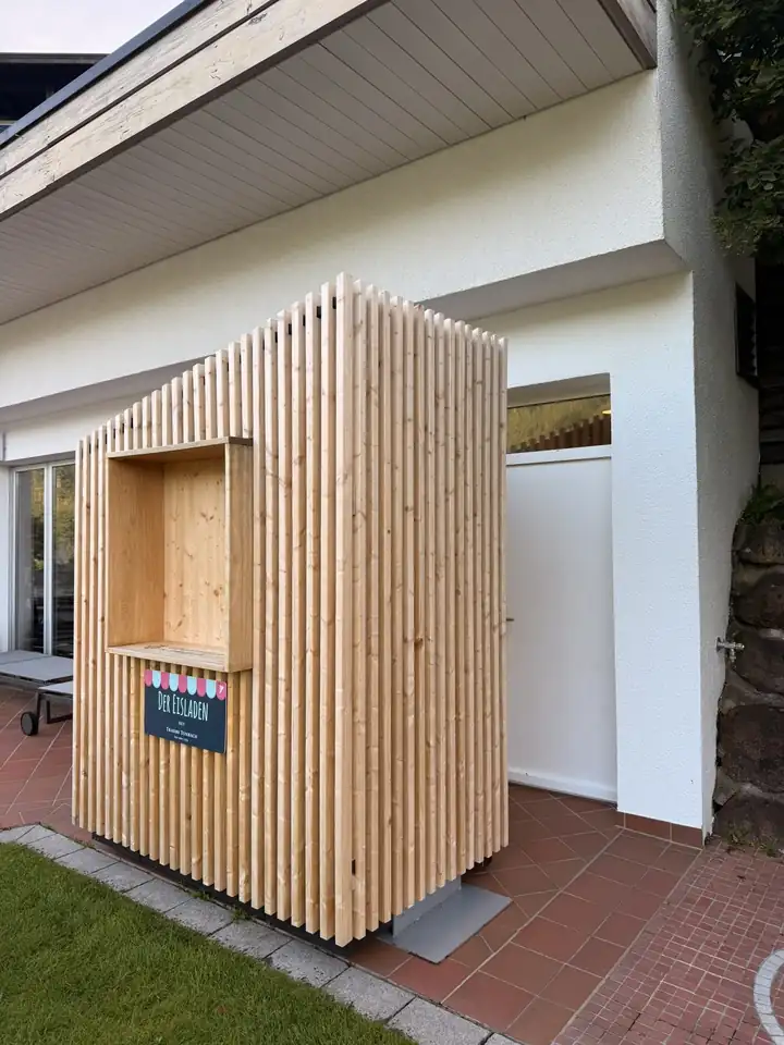 Kleines Häuschen aus hellen Holzlatten mit Verkaufsfenster und dem Schild Der Eisladen auf einer Terrasse mit roten Fliesen