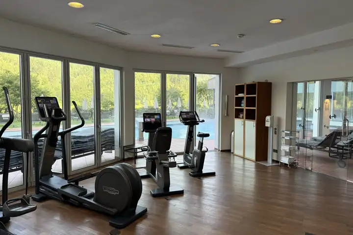Fitnessraum mit Crosstrainern und Ergometern vor einer Glasfront, draußen der Außenpool, Liegen mit Schirmen und ein bewaldeter Hang