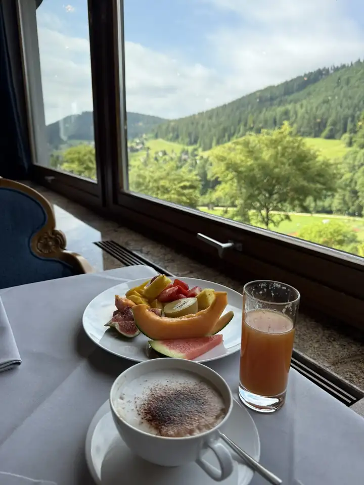 Teller mit Feigen, Papaya, Ananas, Erdbeeren, Kiwi und Melone, daneben ein Cappuccino und ein Glas Saft am Fenster, draußen bewaldete Hänge
