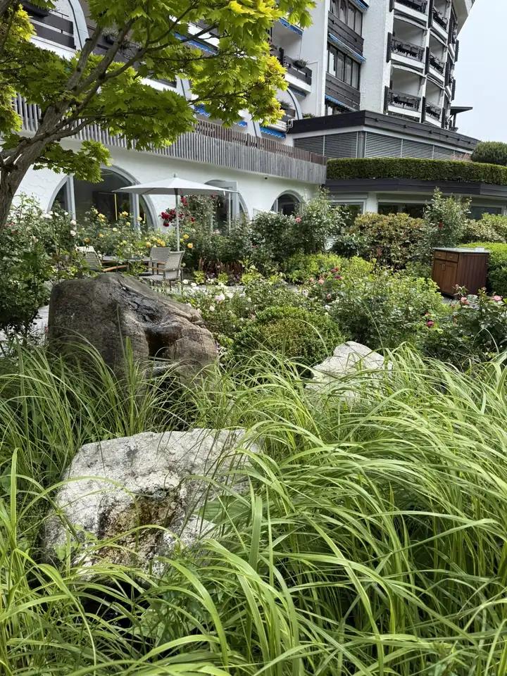 Garten mit Findlingen, hohen Gräsern und Rosen in Rosa, Gelb und Rot vor der Hotelfassade mit Rundbögen, dahinter ein Sonnenschirm über Gartenmöbeln