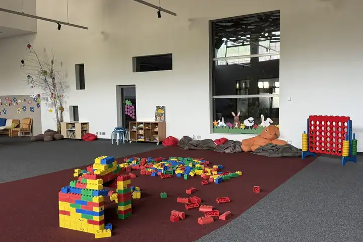 Große helle Spielhalle mit weichen bunten Bausteinen auf einem roten Teppich, Sitzsäcken, einem großen Vier gewinnt Spiel und Regalen mit Büchern