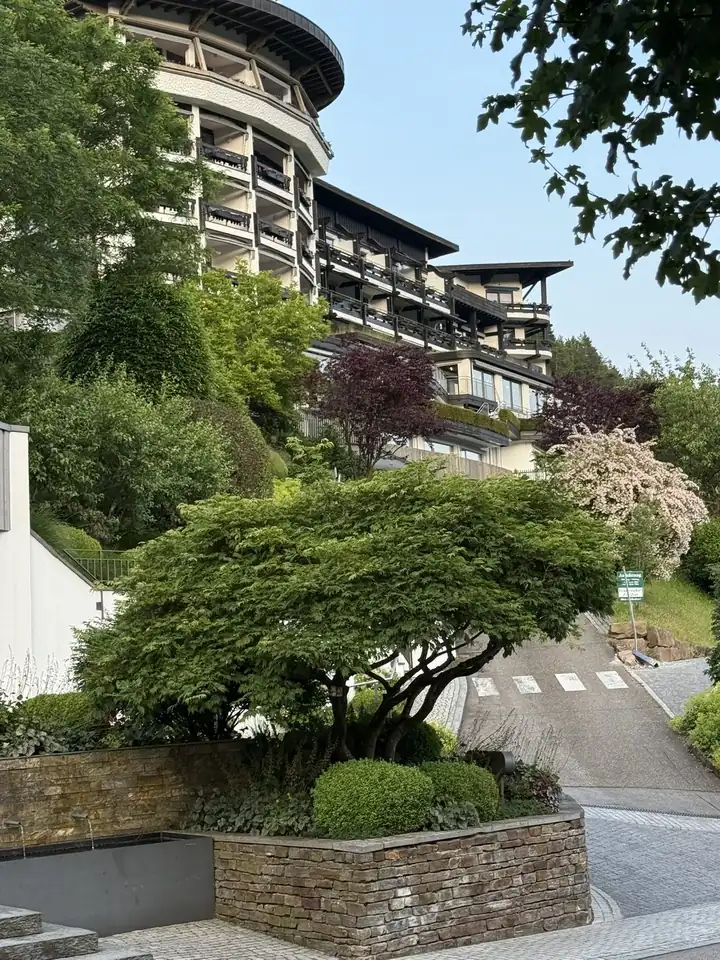 Runder Hotelturm mit dunklen Holzbalkonen über einem Hang mit Büschen, im Vordergrund ein Fächerahorn auf einer Natursteinmauer und eine ansteigende Zufahrt