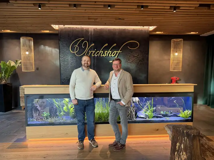 Benjamin Brandl im hellen Pullover und Lars Laschinski im beigen Sakko lehnen am Empfangstresen des Ulrichshof, darunter ein beleuchtetes Aquarium, dahinter der goldene Schriftzug