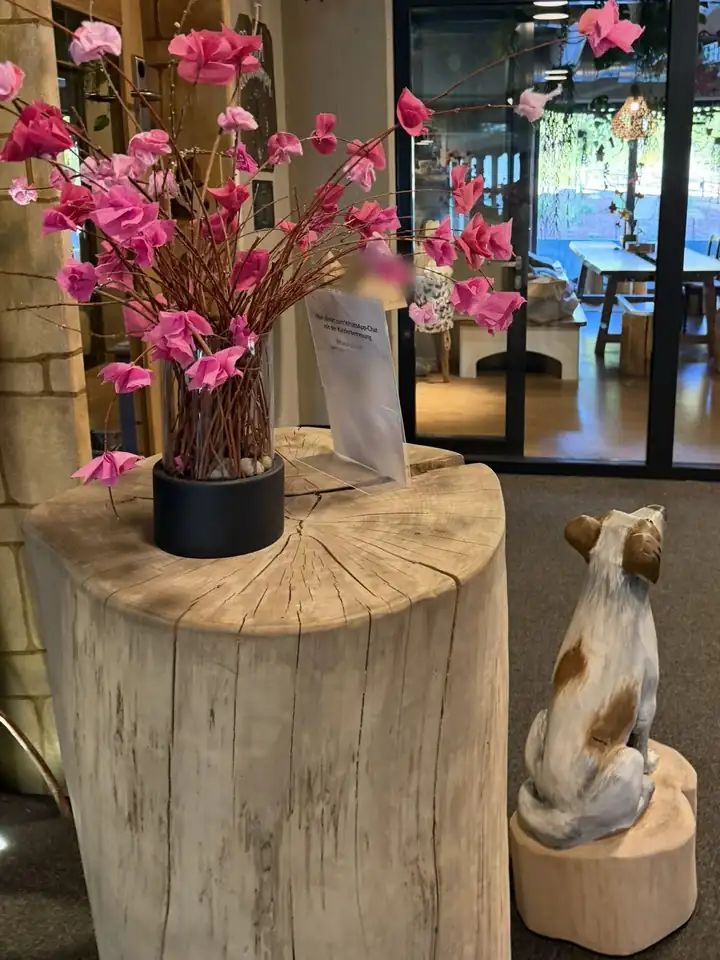 Baumstumpf mit Vase voller rosa Papierblüten neben einer geschnitzten Hundefigur aus Holz, dahinter ein Raum mit langer Tafel