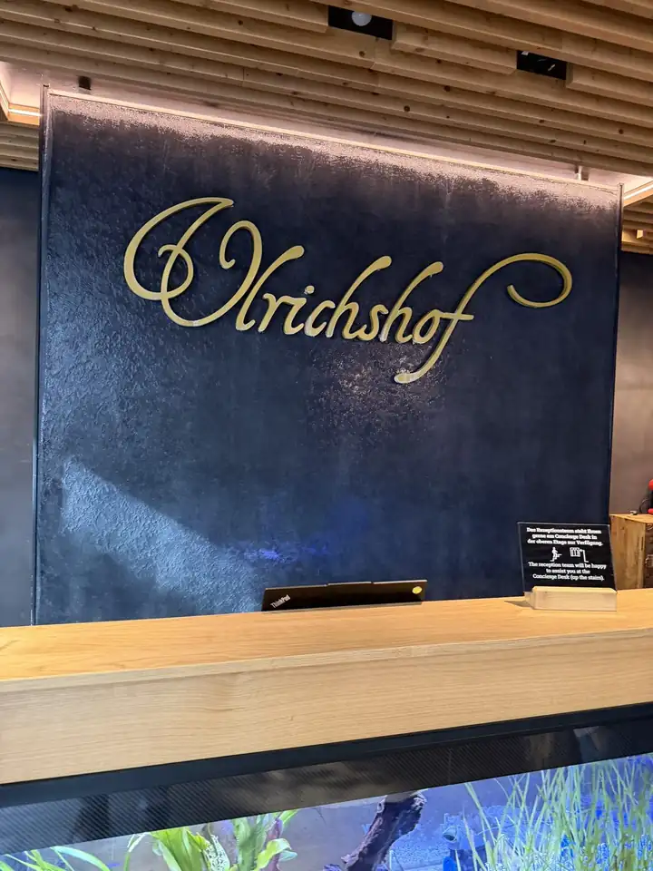 Rezeption des Ulrichshof mit goldenem Schriftzug auf dunkler Wand, darunter ein Tresen aus hellem Holz und ein beleuchtetes Aquarium