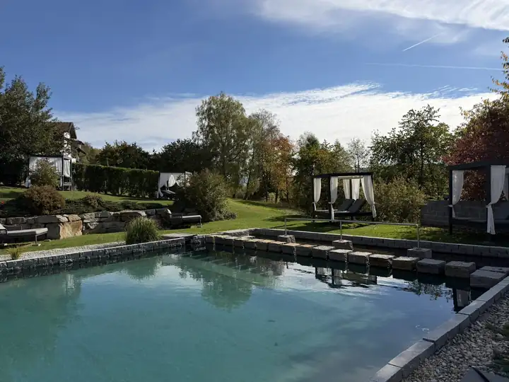 Naturpool mit Trittsteinen und Geländer, dahinter ein Garten mit Himmelbetten, weißen Vorhängen, Laubbäumen und Natursteinmauer