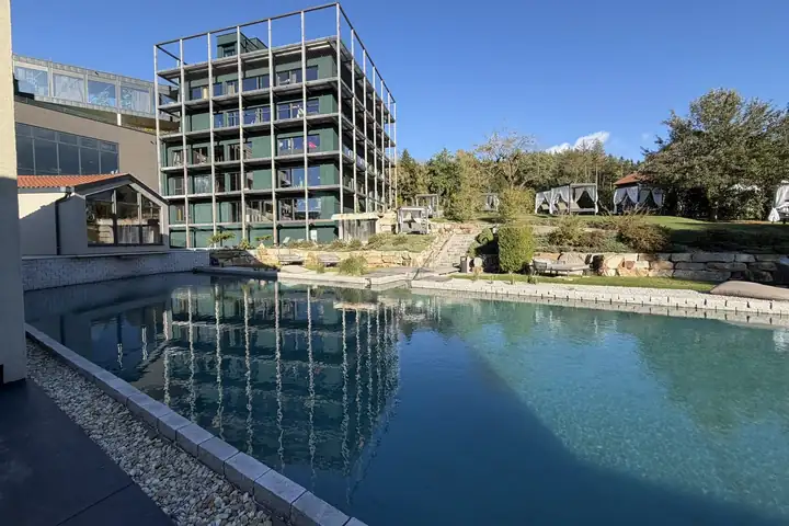 Neubau mit Stahlrahmen und dunkelgrünen Balkonen, der sich in einem Naturpool spiegelt, daneben Himmelbetten auf dem Rasen unter blauem Himmel