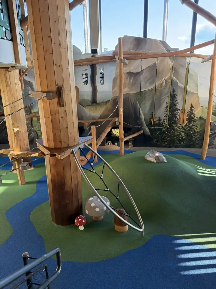Kletterwald in einer Halle mit dicken Holzstämmen, Netzen, einem Ring zum Klettern und Pilzen aus Holz auf grünem Boden