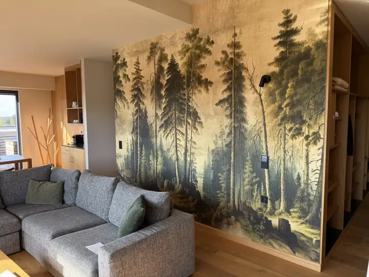 Wohnraum der Suite mit grauem Ecksofa vor einer Tapete mit Waldgemälde, rechts offene Schränke aus Eiche