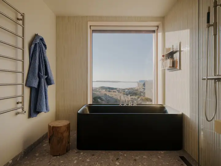 Bad mit schwarzer Badewanne vor einem Fenster mit Meerblick, daneben Handtuchheizkörper und ein blauer Bademantel