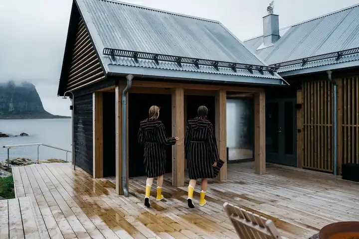 Zwei Gäste in gestreiften Bademänteln und gelben Gummistiefeln gehen über eine nasse Holzterrasse zum Saunahaus