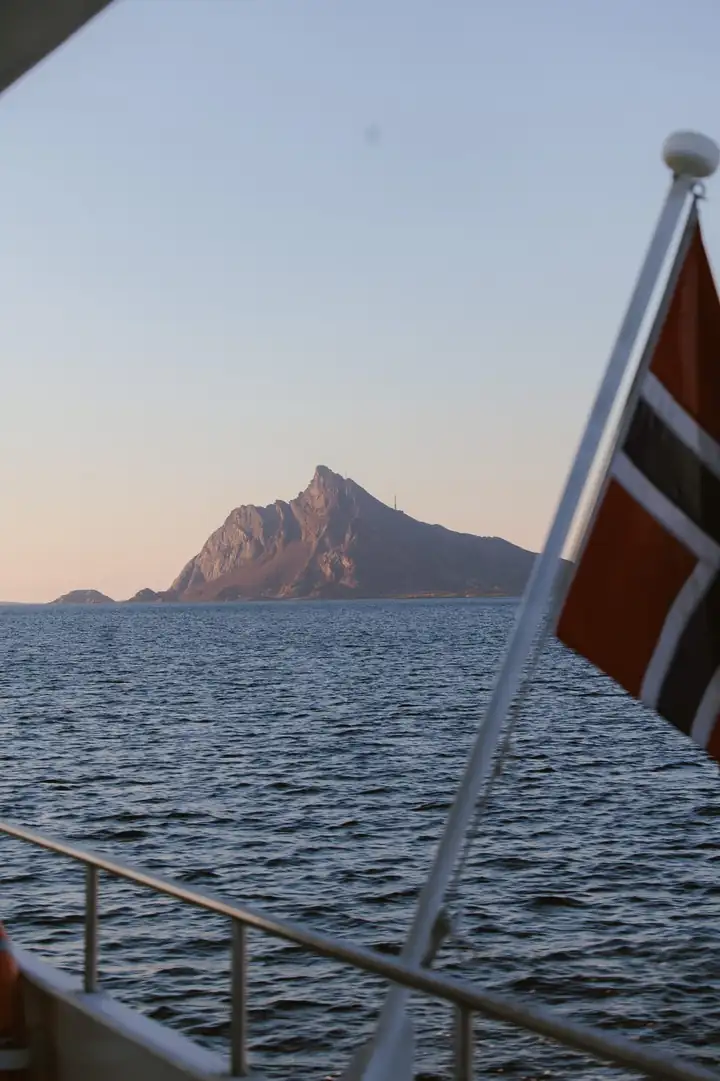 Blick vom Deck eines Schiffs mit norwegischer Flagge auf einen spitzen Berg über dem Meer
