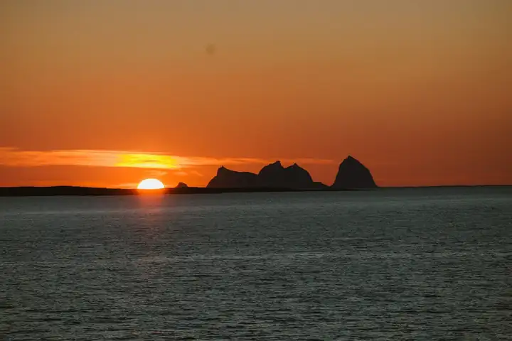 Orangefarbener Abendhimmel, die Sonne steht knapp über dem Meer neben den Umrissen felsiger Inseln