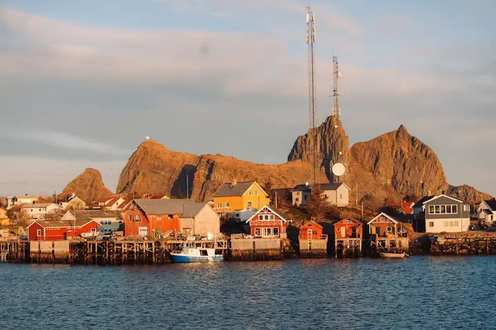 Bunte Holzhäuser und Bootshäuser am Hafen von Træna unter einem Felsrücken mit Funkmasten im Abendlicht